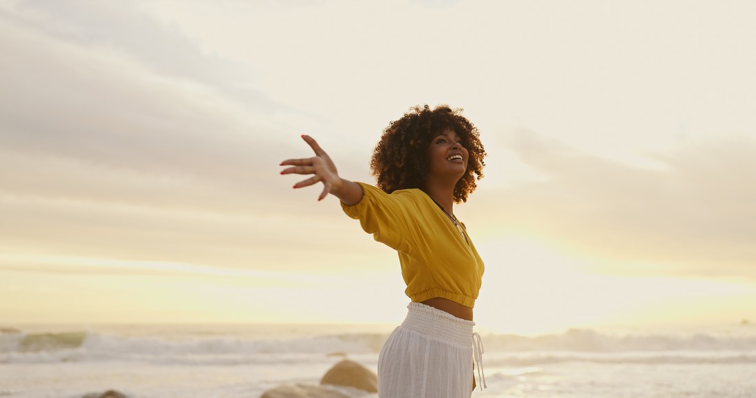 A woman on the beach at sunset with her arms open to the sky — vitality and wellbeing.