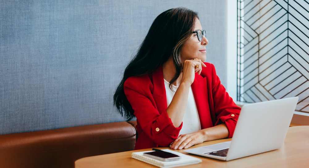 A businesswoman in a red blazer at her laptop, looking thoughtfully out an office window — planning a corporate wellness programme.