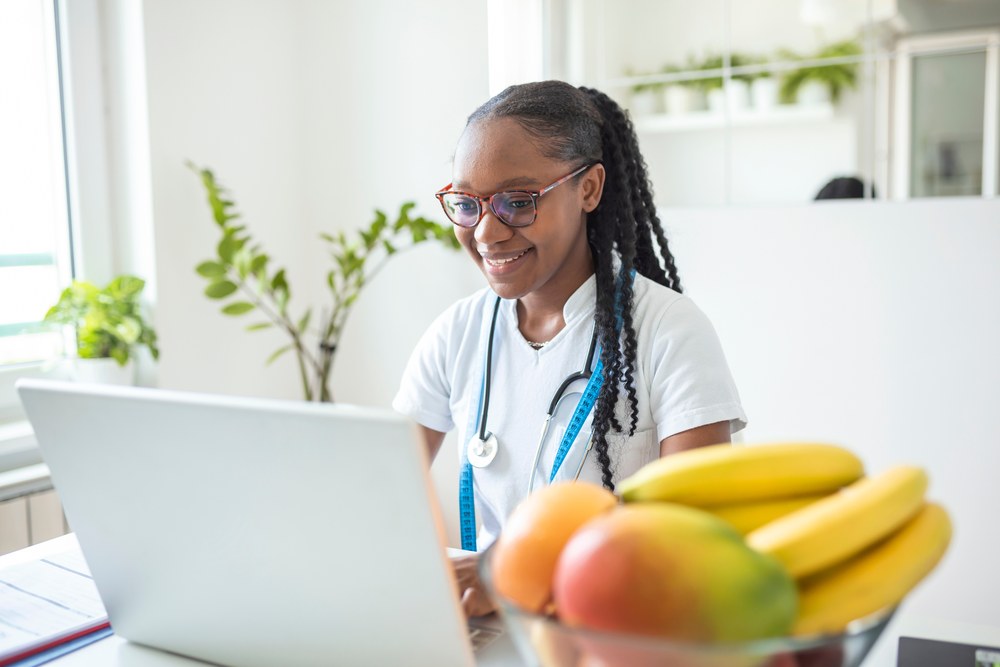 A nutritionist wearing a stethoscope reviewing results on a laptop, a bowl of fresh fruit on the desk.
